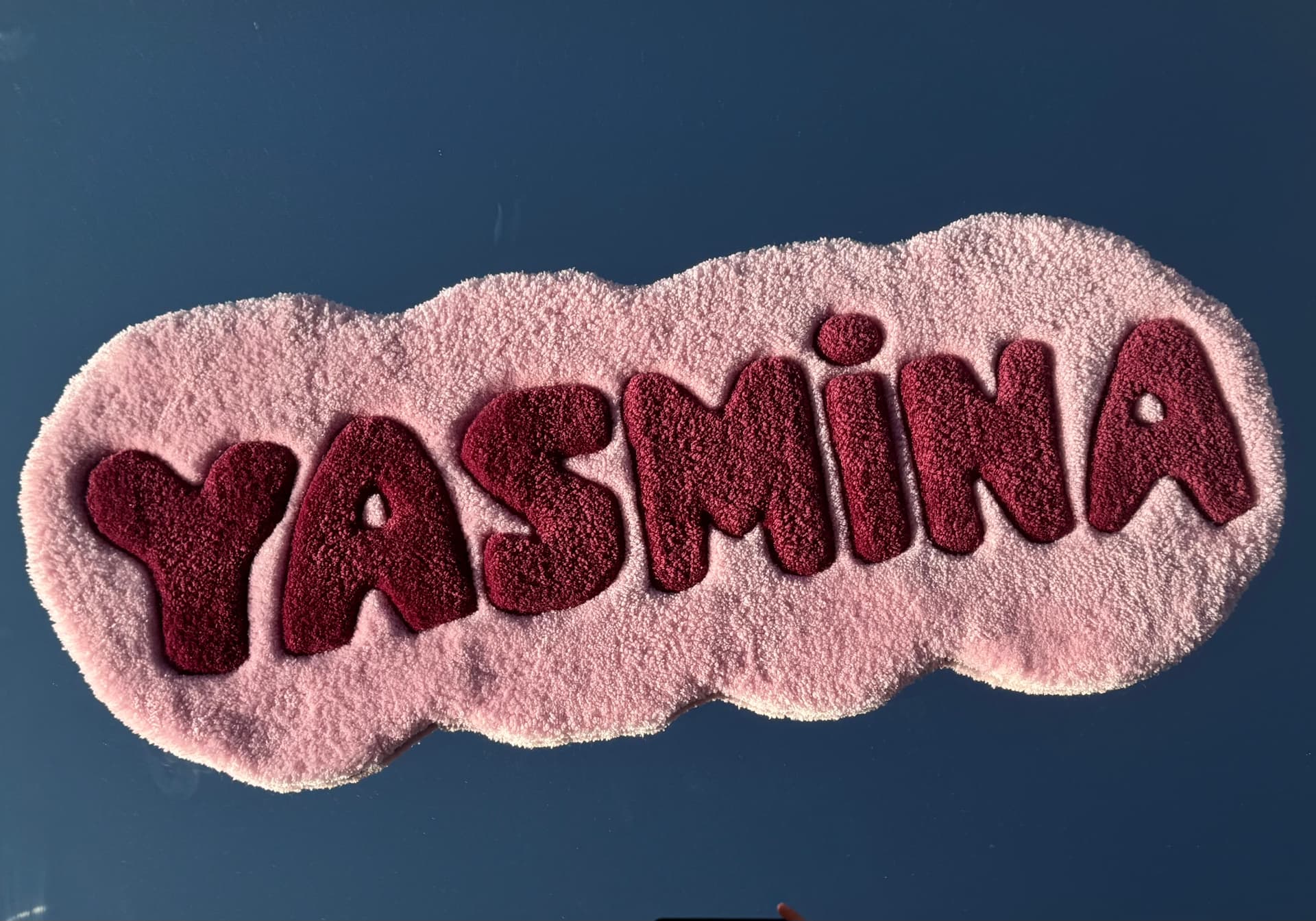 This is a custom hand-tufted name rug featuring the name “YASMINA” in bold, playful lettering. The design uses a soft blush-pink background with deep burgundy letters, creating a beautiful contrast. The letters are carefully carved around the edges, giving them a raised, 3D effect and making the name stand out. The rug has a fun organic, wavy silhouette that follows the shape of the lettering rather than a traditional square or circle.