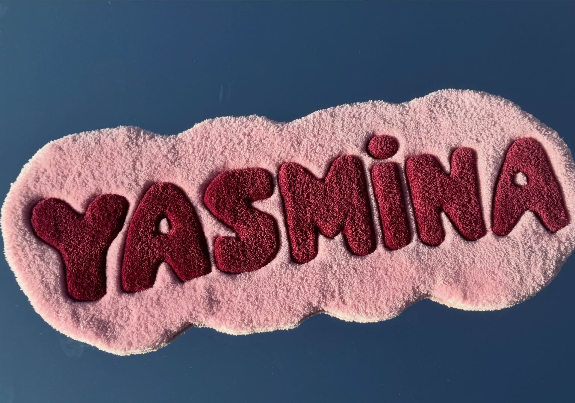This is a custom hand-tufted name rug featuring the name “YASMINA” in bold, playful lettering. The design uses a soft blush-pink background with deep burgundy letters, creating a beautiful contrast. The letters are carefully carved around the edges, giving them a raised, 3D effect and making the name stand out. The rug has a fun organic, wavy silhouette that follows the shape of the lettering rather than a traditional square or circle.